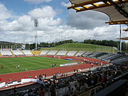 Interior of a sports stadium. There is a running track surrounding a central grassed area. In the distance there are the outer stands. In the foreground is the main stand, with plenty of people seated