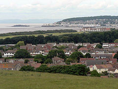 Multiple houses and other buildings around a bay into which a pier projects. On the background are hills