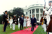 Two couples walk down a red carpet, away from the White House. Military Honor Guard from all services stand at attention along both sides of the carpet.