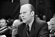 A man in a suit is seated at a table as he speaks into a bank of microphones. An audience is visible behind him.