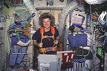 A woman running on a treadmill, anchored by orange straps. The wall behind her has a variety of items including clothing, ventilation hoses and instrument panels affixed to it.