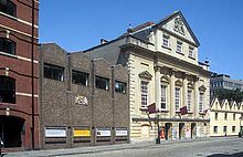 An imposing eighteenth-century building with three entrance archways, large first-floor windows and an ornate peaked gable end above. On the left a twentieth-century grey brick building with a gilded crest and on the right a cream-coloured building with four pitched roofs. In front a cobbled street.