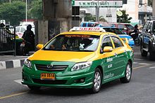 A Toyota saloon/sedan driving on the road, its top half painted in yellow and the bottom half in green, with a sign on the roof saying 