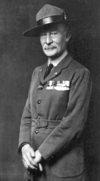 Black and white photograph of a man in military uniform, with his medals pinned to the left side of his jacket. He is wearing a wide-brimmed hat and holding a walking stick in both hands.