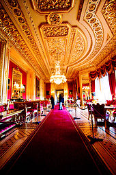 A photograph of a large room with a long red carpet stretching through the middle of it and windows on the right hand side. Furniture fills both sides of the room. The ceiling contains ornate plasterwork and a chandelier hangs down from the middle of the picture.