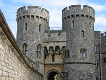 A photograph of a stone gatehouse, with two large, circular towers on each side of the gateway dominating the picture. A stone wall stretches alongside the left hand side of the picture.