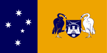 Left third: White stars of the Southern Cross on blue field.  Right two-thirds; black and white swans holding a blue shield with a white castle above a white flower and below a sword and scepter arranged in an 