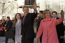 Same teenage girl, man and woman walk down a broad street in wintertime, as security personnel trail and a crowd looks on