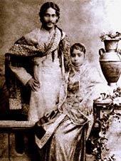 Black-and-white photograph of a finely dressed man and woman: the man, smiling, stands akimbo behind a settle with a shawl draped over his shoulders and in Bengali formal wear. The woman, seated on the settle, is in elaborate dress and shawl; she leans against a carved table supporting a vase and flowing leaves.