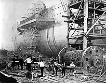 An old photograph showing a large iron paddlewheel ship being launched sideways, with workmen thrusting large baulks of timber under a large drum of iron chains