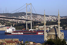 A large ship passes underneath a suspension bridge and behind a small mosque with two towering minarets.