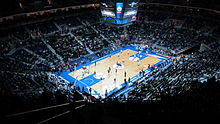 A brightly lit basketball court and game, with spectators filling the surrounding darkened bowl of seats