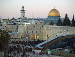 A large open area with hundreds of people, bounded by old stone walls. Beyond it are houses and a few trees, to the right is a mosque with large golden dome, and to the left – a minaret.